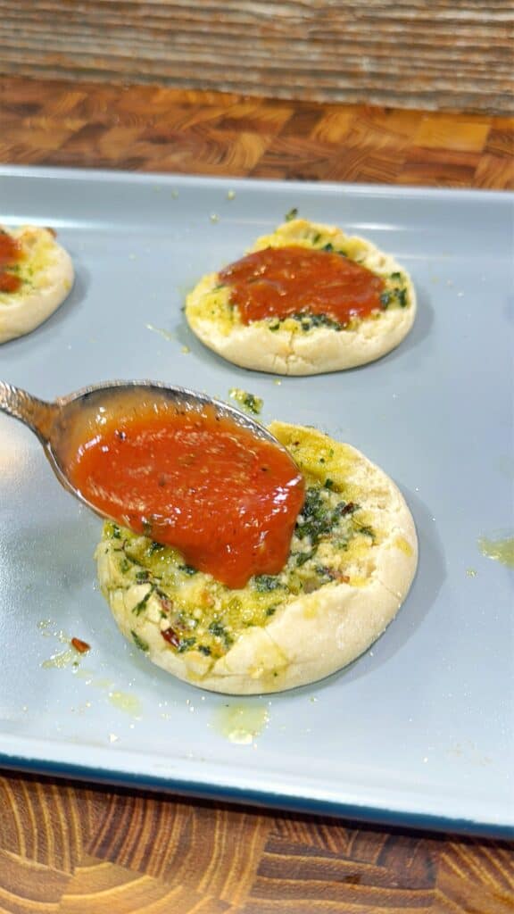 A spoon spreading tomato sauce onto an English muffin topped with a spinach and cheese mixture, placed on a baking sheet.