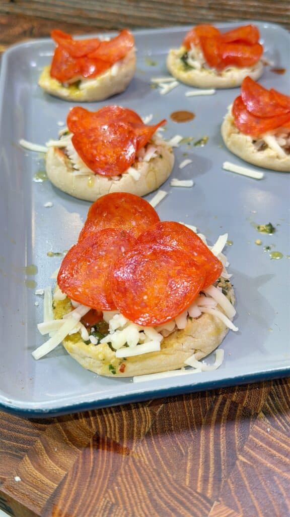 Mini pizzas with mozzarella cheese, pepperoni slices, and herbs are arranged on a baking tray, ready to be baked. The tray is on a wooden surface.