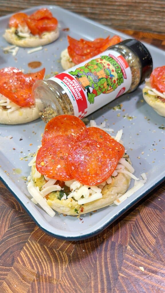 A baking tray holds mini pizzas on English muffins topped with cheese, spinach, and pepperoni slices. A bottle of Italian seasoning, featuring cartoon turtles on the label, is tipped over, sprinkling spices on the pizzas.