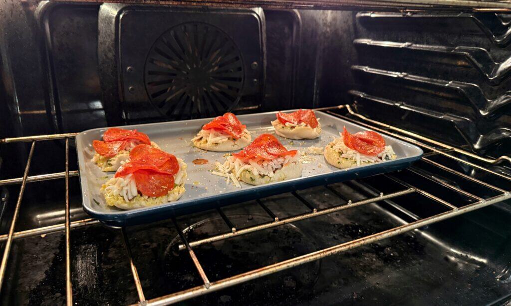 A baking tray with mini pizzas topped with cheese and pepperoni is on the middle rack of an oven, in the process of baking.