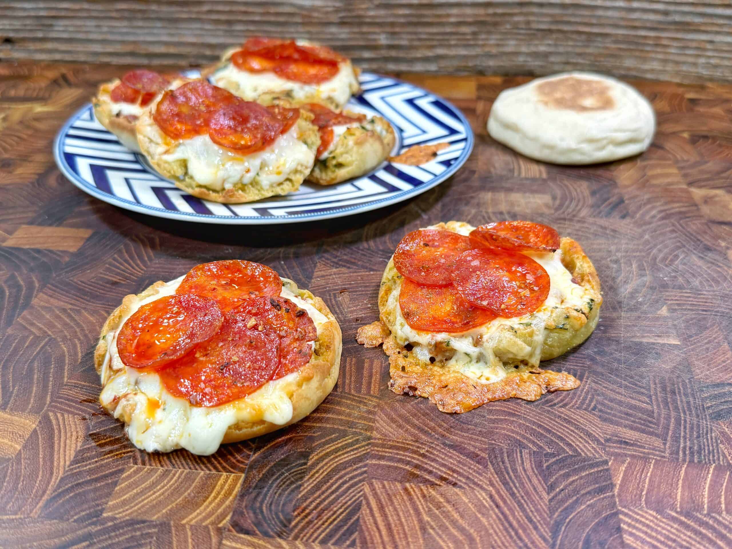 Mini pepperoni pizzas with melted cheese on English muffin halves are on a chevron-patterned plate and a wooden surface. A plain English muffin is in the background.