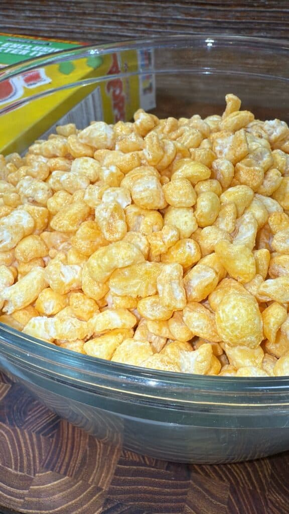 A clear glass bowl filled with golden, puffy cereal pieces sits on a wooden surface. In the background, part of a colorful cereal box is visible. This is for a homemade beaver nuggets recipe.
