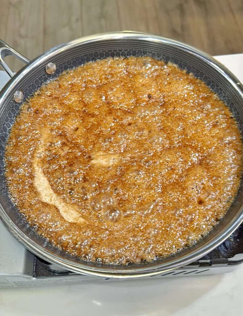 A close-up of a pan on a stovetop with bubbling brown liquid, likely sugar caramelizing or syrup boiling, with a metal handle visible on the left. The background shows a wooden floor.