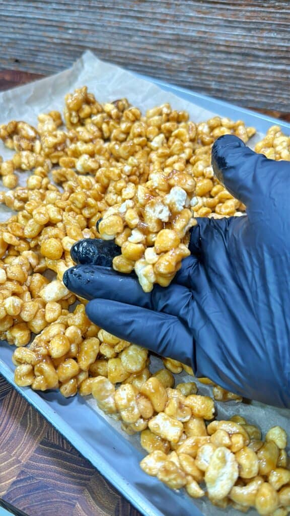 A gloved hand holds a cluster of caramel-coated popcorn above a tray lined with parchment paper and filled with more caramel popcorn.