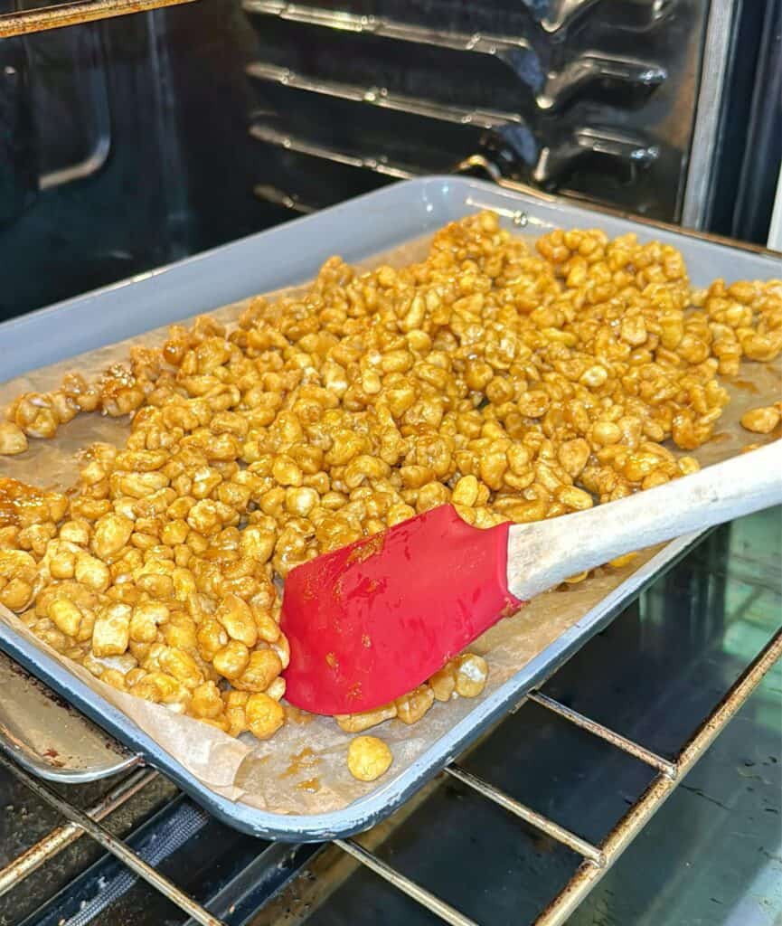 A baking tray lined with parchment paper holds a layer of caramel-coated popcorn being stirred with a red silicone spatula inside an oven.