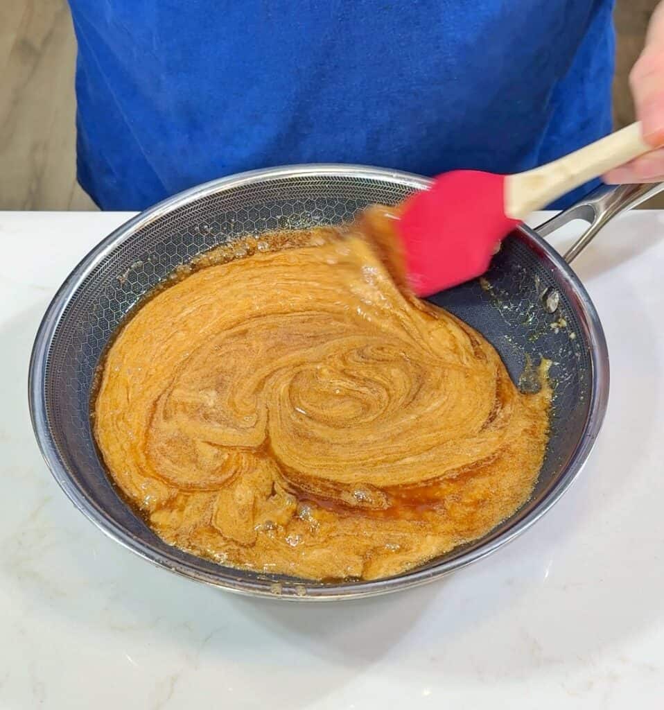 A person in a blue shirt stirs a thick, caramel-colored mixture in a frying pan with a red spatula on a white countertop.
