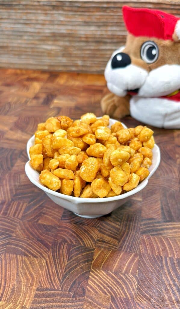 A white bowl filled with crunchy, golden caramel popcorn sits on a wooden surface. In the background, a plush toy dog with a red hat and scarf is partially visible.