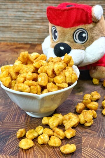 A small white bowl filled with caramel popcorn sits on a wooden surface. Pieces of popcorn are scattered beside it. A plush toy wearing a red outfit and hat is positioned behind the bowl, facing forward.