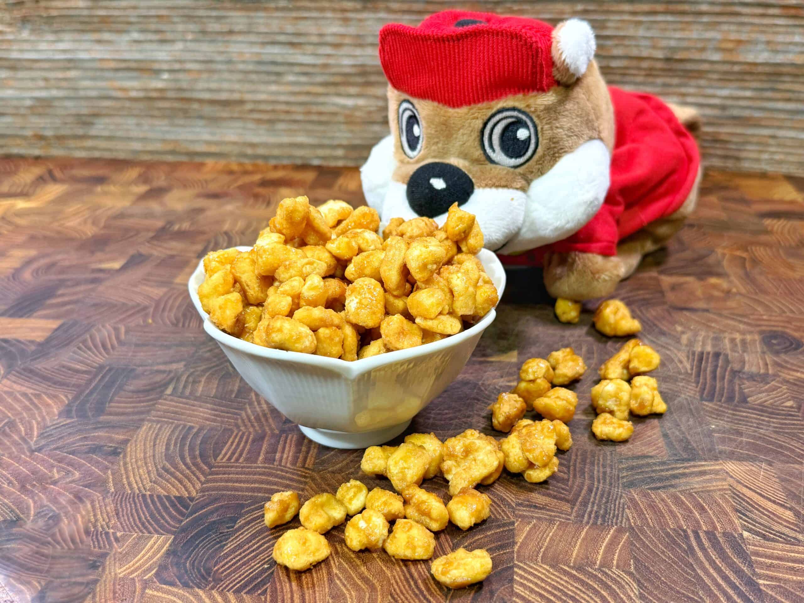 A small white bowl filled with caramel popcorn sits on a wooden surface. Pieces of popcorn are scattered beside it. A plush toy wearing a red outfit and hat is positioned behind the bowl, facing forward.