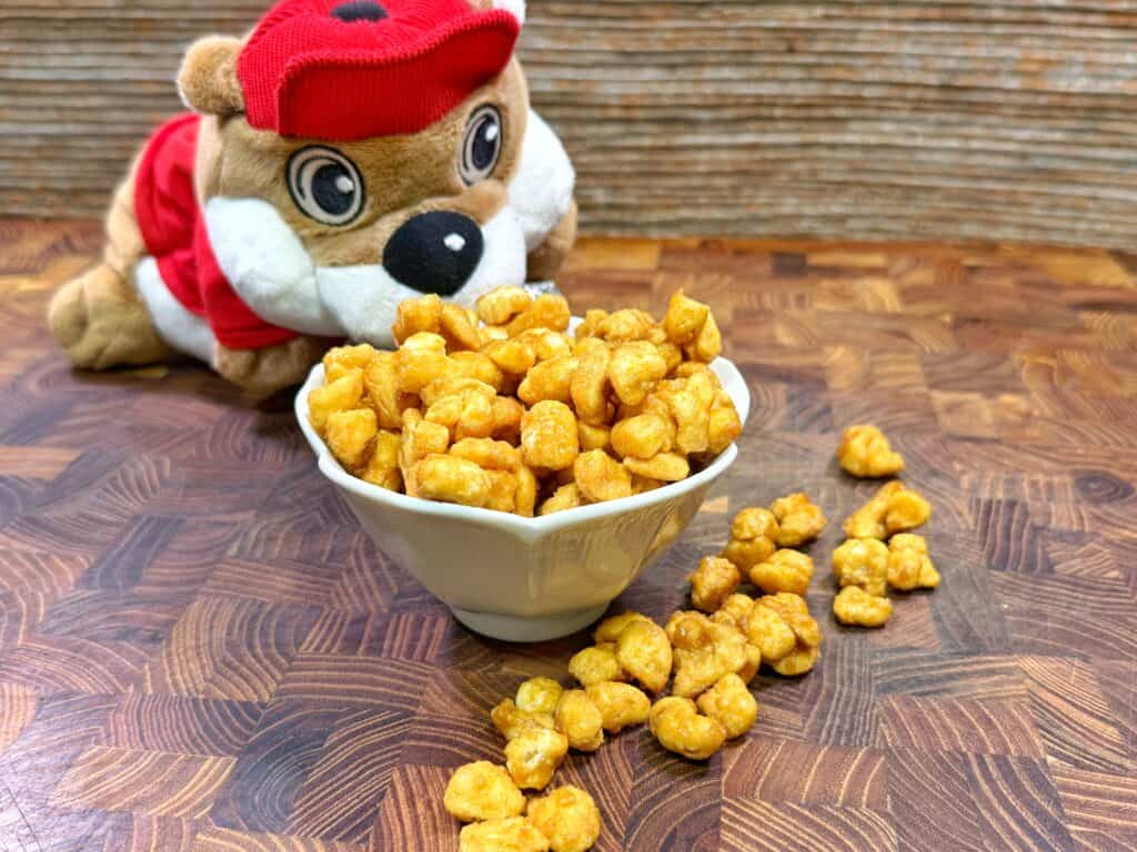 A plush toy chipmunk in a red hat and vest sits behind a white bowl filled with caramel-coated popcorn, with some popcorn pieces spilled onto a wooden surface.