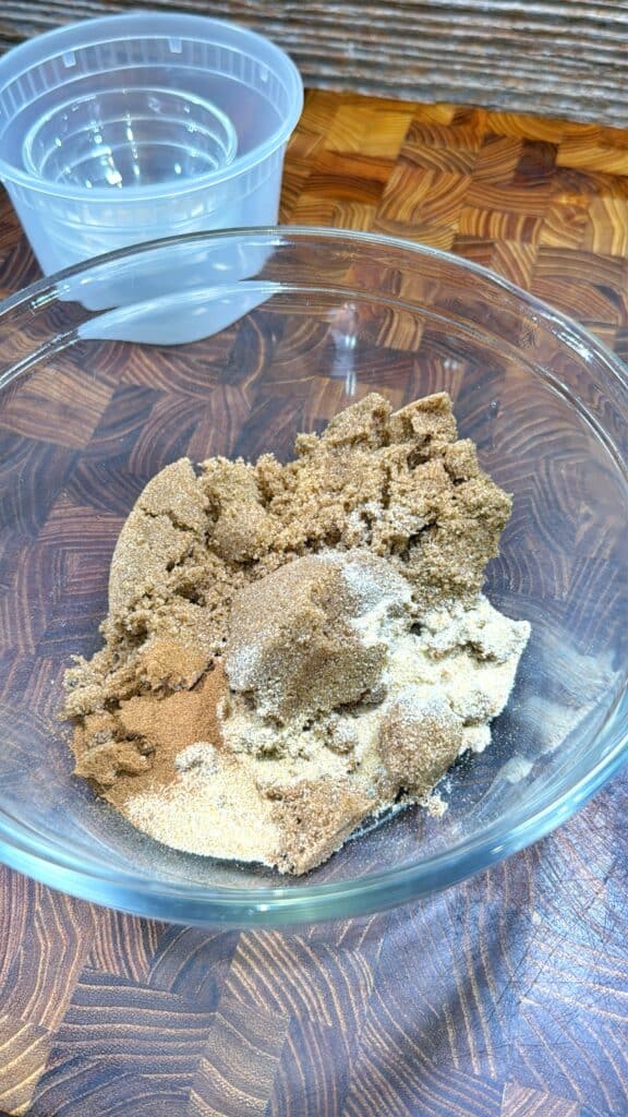 A glass bowl on a wooden surface contains brown sugar and various ground spices. In the background, there is a small empty plastic container.