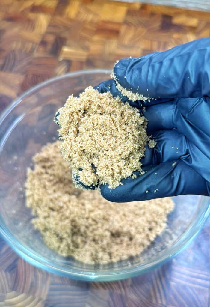 A hand in a black glove holds a clump of light brown sugar above a glass bowl filled with more brown sugar, all on a wooden surface.