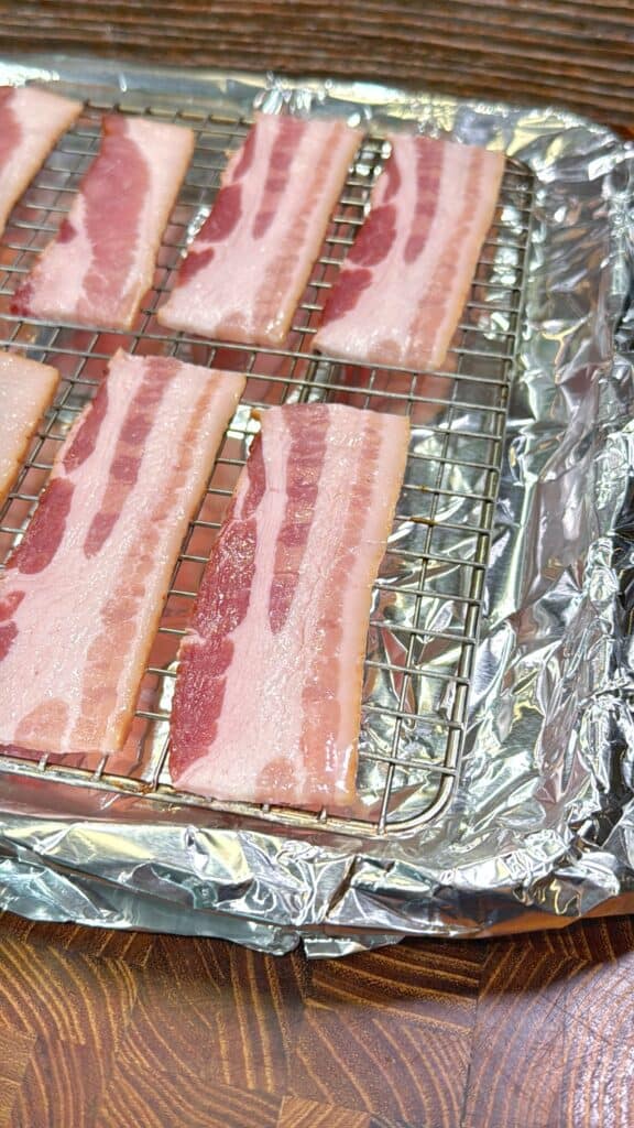 Raw strips of bacon are laid out on a metal rack placed over a baking sheet lined with aluminum foil, ready to be cooked. The setup rests on a wooden surface.
