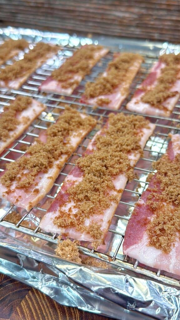 Raw bacon strips on a wire rack over foil, topped with brown sugar, ready to be cooked.