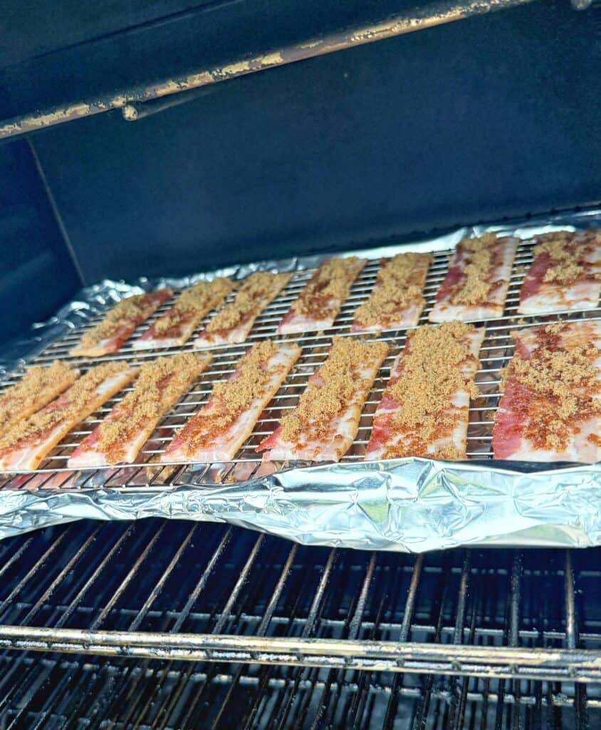 Strips of bacon topped with brown sugar are arranged on a wire rack over a foil-lined baking sheet, placed inside an oven for cooking.