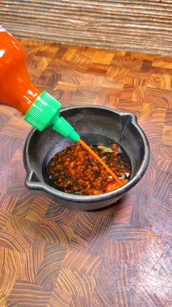 A bottle with a green cap pours bright orange hot sauce into a small black bowl filled with a dark, seasoned liquid, on a wooden surface.