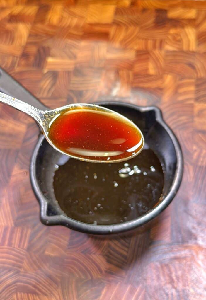 A spoon holds a small amount of brown liquid over a black bowl filled with more of the same liquid, set on a wooden surface.