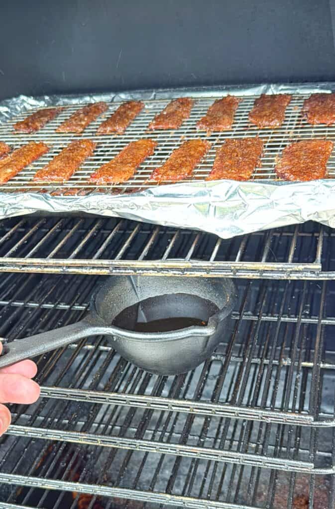 A hand holds a small black pan with liquid beneath a grill rack of seasoned meat strips on foil inside a smoker or oven.