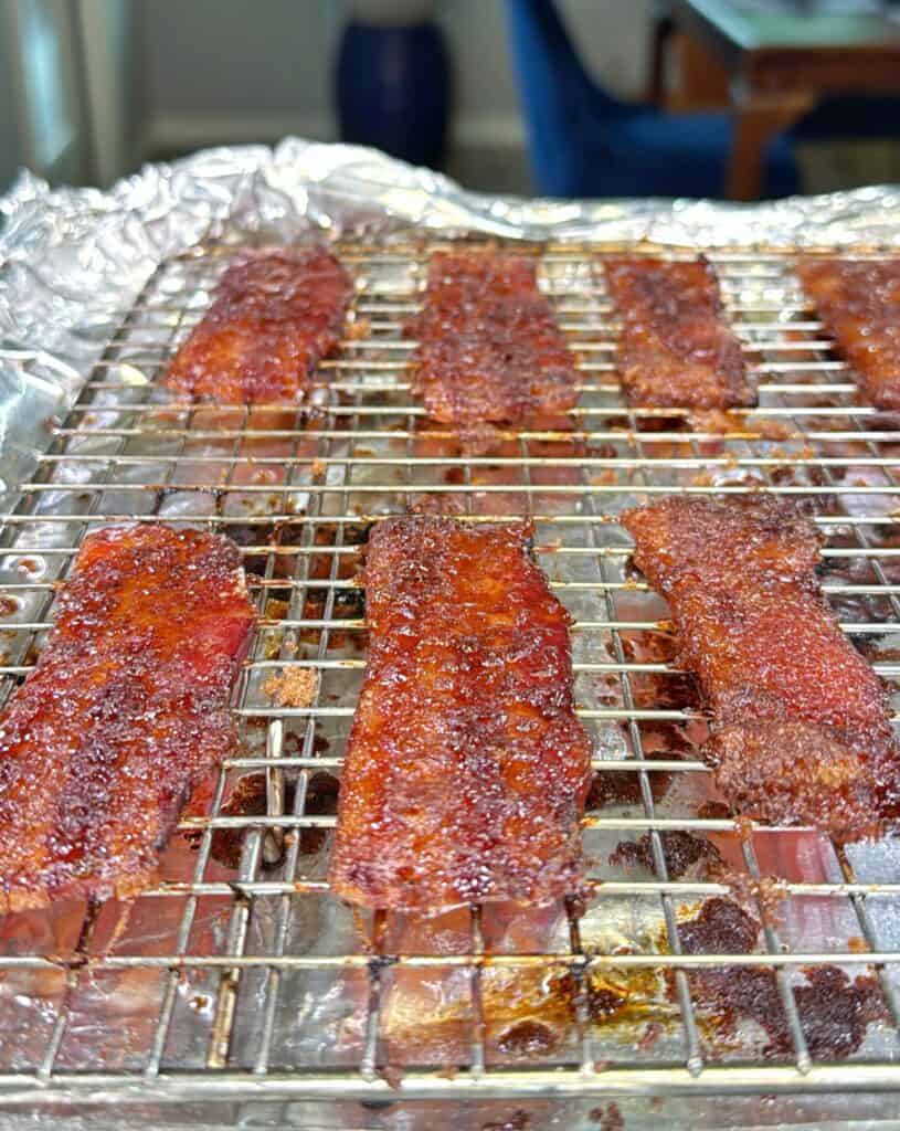 Several strips of crispy glazed bacon are cooling on a wire rack over a foil-lined baking sheet. The bacon appears caramelized and well-cooked, with a rich, brown color.