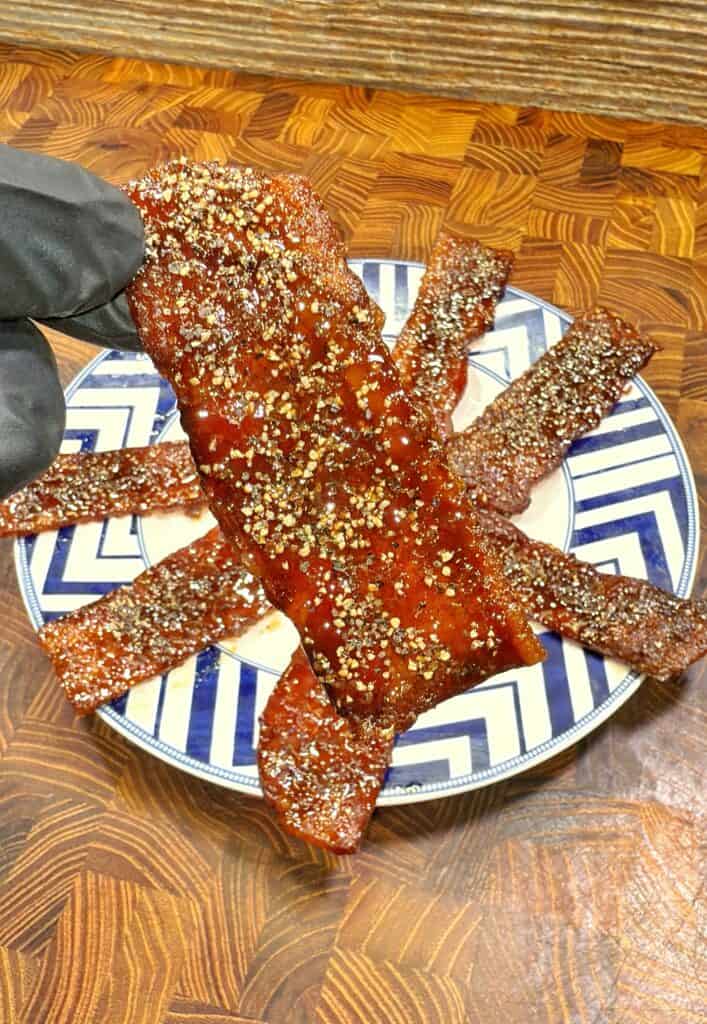 A gloved hand holds a piece of thick, glazed bacon coated with cracked black pepper above a plate with several similar bacon strips arranged in a sunburst pattern on a wooden surface. Recipe is for maple sriracha candied bacon