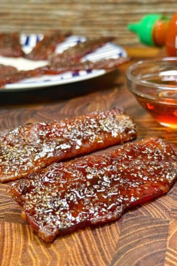 Two pieces of glazed, peppered beef jerky are on a wooden surface, with a small bowl of amber liquid and a bottle of Sriracha sauce nearby. A plate with more jerky is in the background.