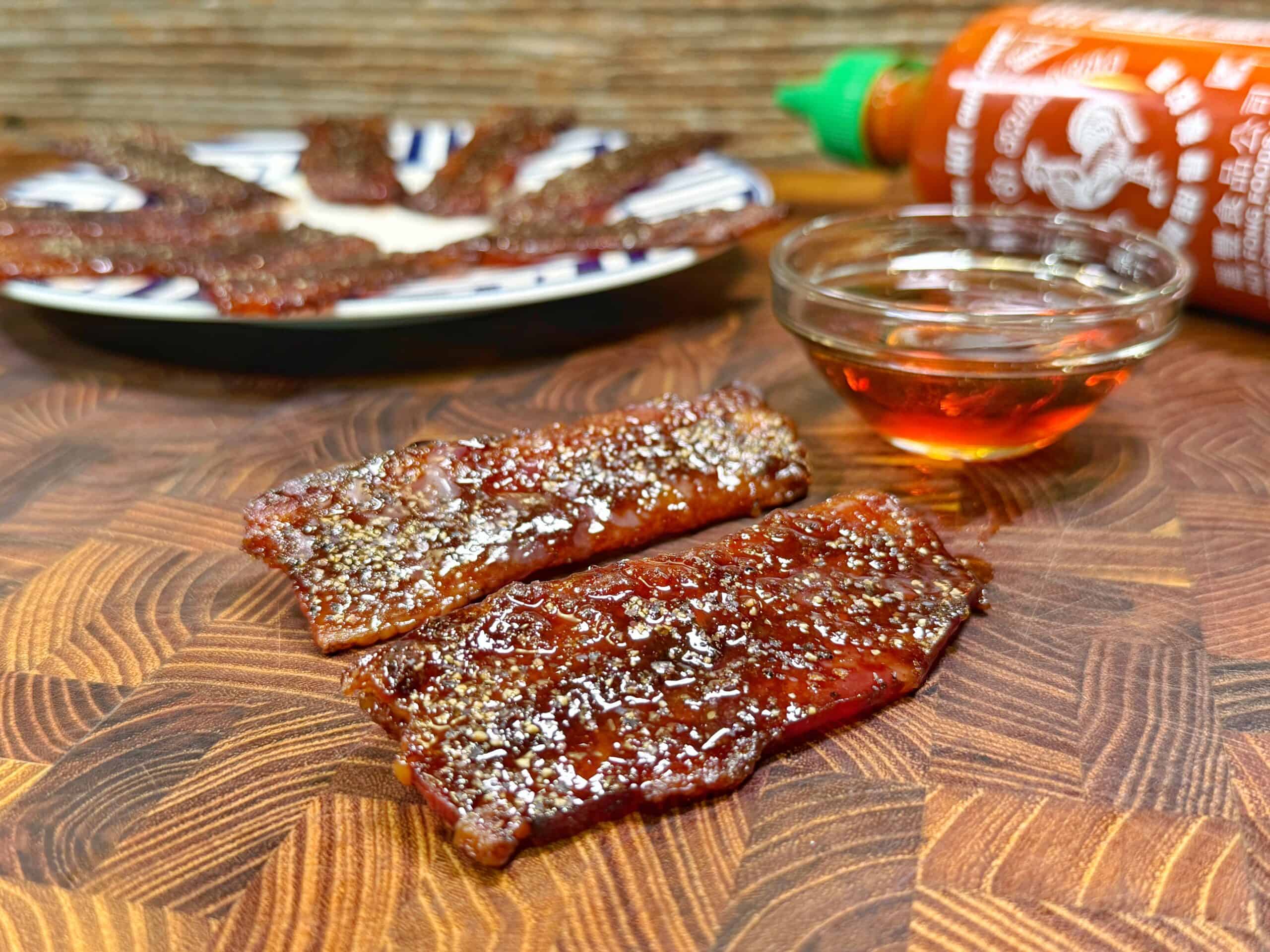 Two pieces of glazed, peppered beef jerky are on a wooden surface, with a small bowl of amber liquid and a bottle of Sriracha sauce nearby. A plate with more jerky is in the background.