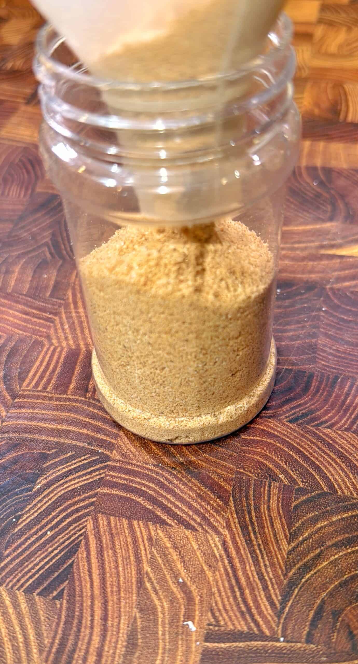A close-up of a glass jar being filled with light brown granulated sugar through a paper funnel, placed on a wooden surface with a geometric pattern.