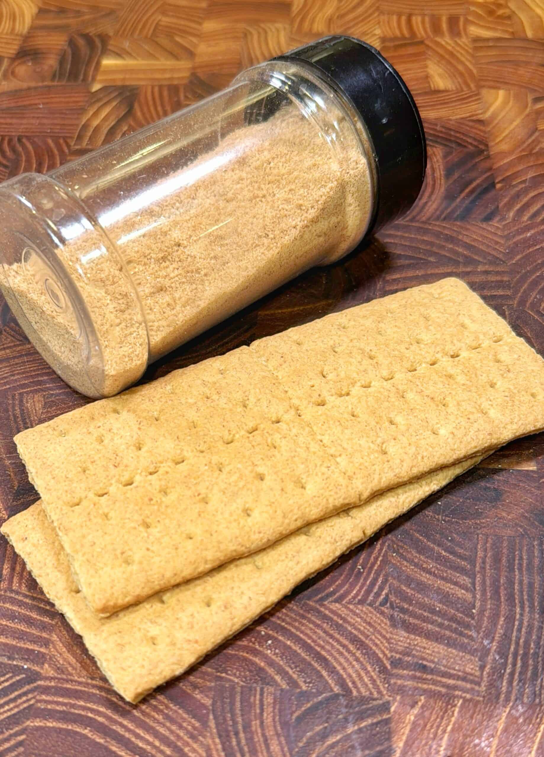 A jar of graham cracker crumbs lies on its side next to two whole graham cracker sheets on a wooden surface.