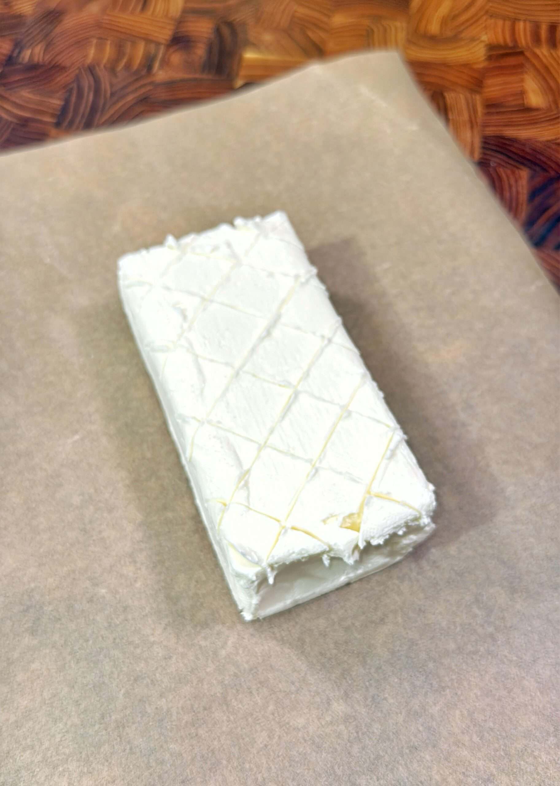 A rectangular block of cream cheese with a diamond pattern on top sits on a piece of parchment paper, on a wooden surface.