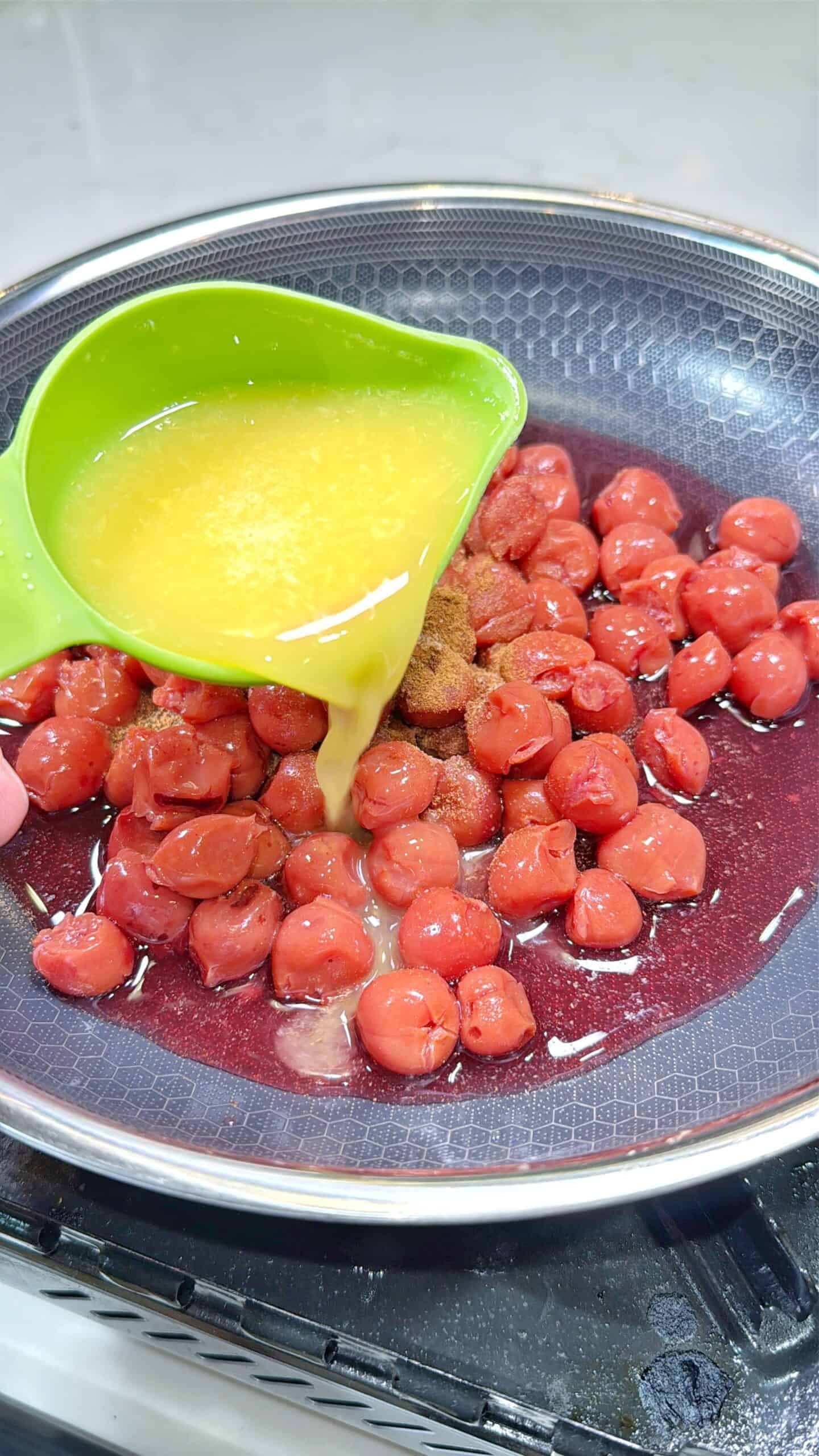 A green bowl pours melted butter over sliced red sausages cooking in a frying pan on a stove.