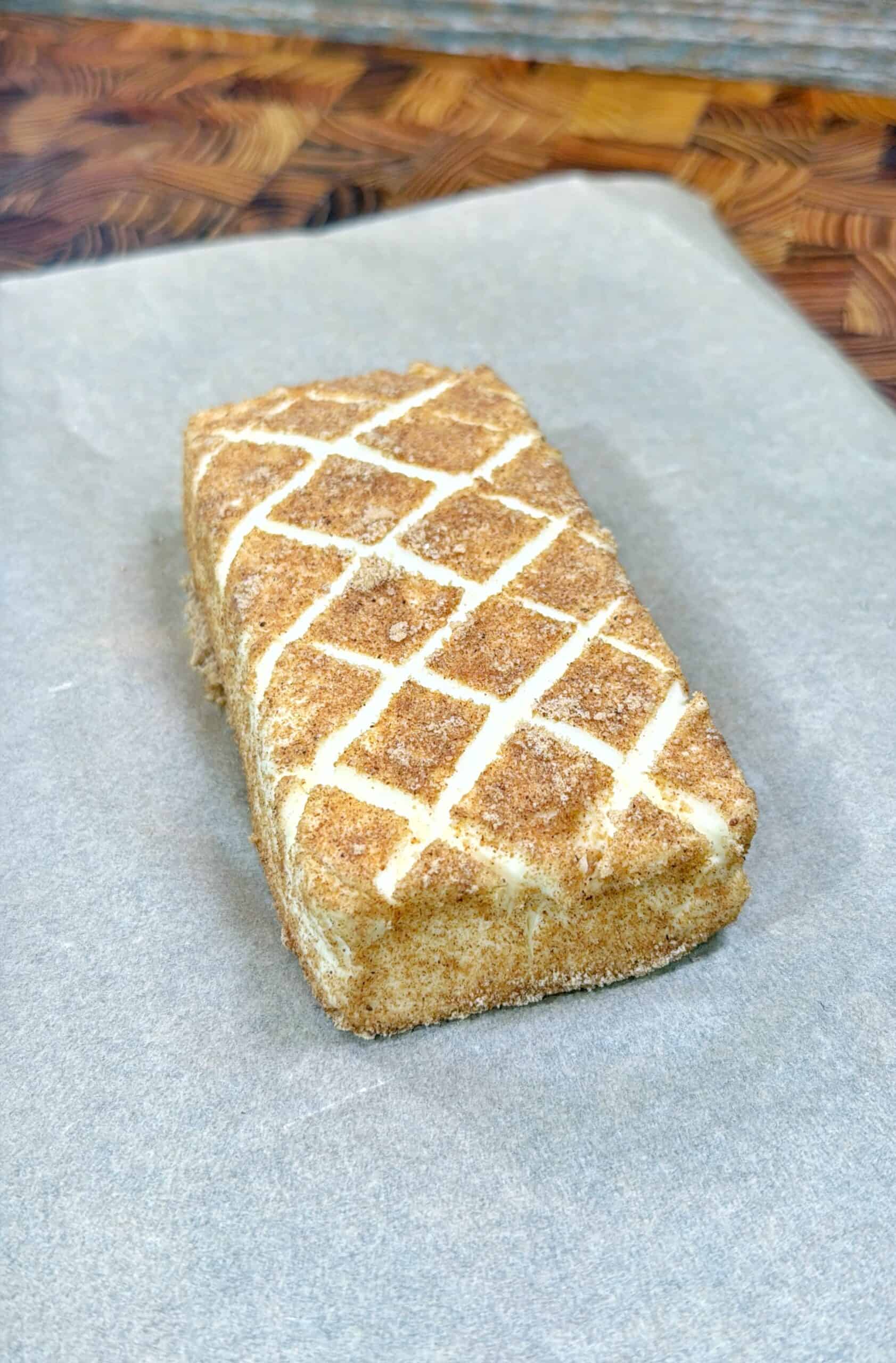 A rectangular block of butter, coated in cinnamon and sugar with a crisscross pattern on top, sits on a sheet of parchment paper on a wooden surface.