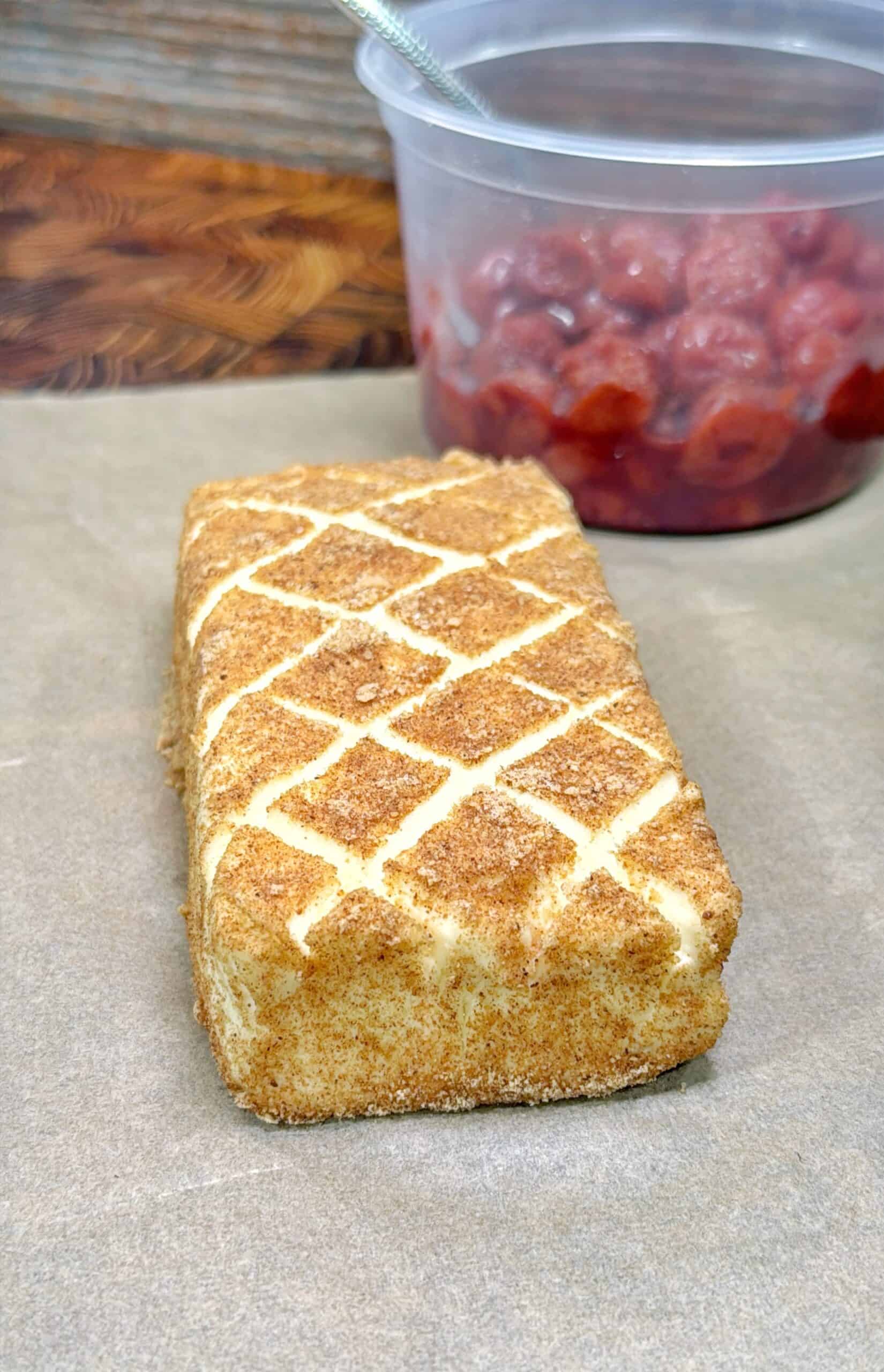 A rectangular cake with a crisscross pattern on top sits on parchment paper. In the background, there is a clear container filled with red fruit, possibly cherries, with a spoon inside.