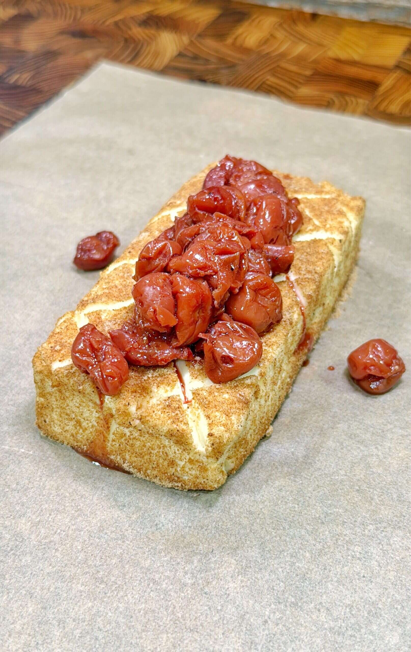 A rectangular loaf of cake topped with glossy, red cherries sits on parchment paper on a wooden surface.