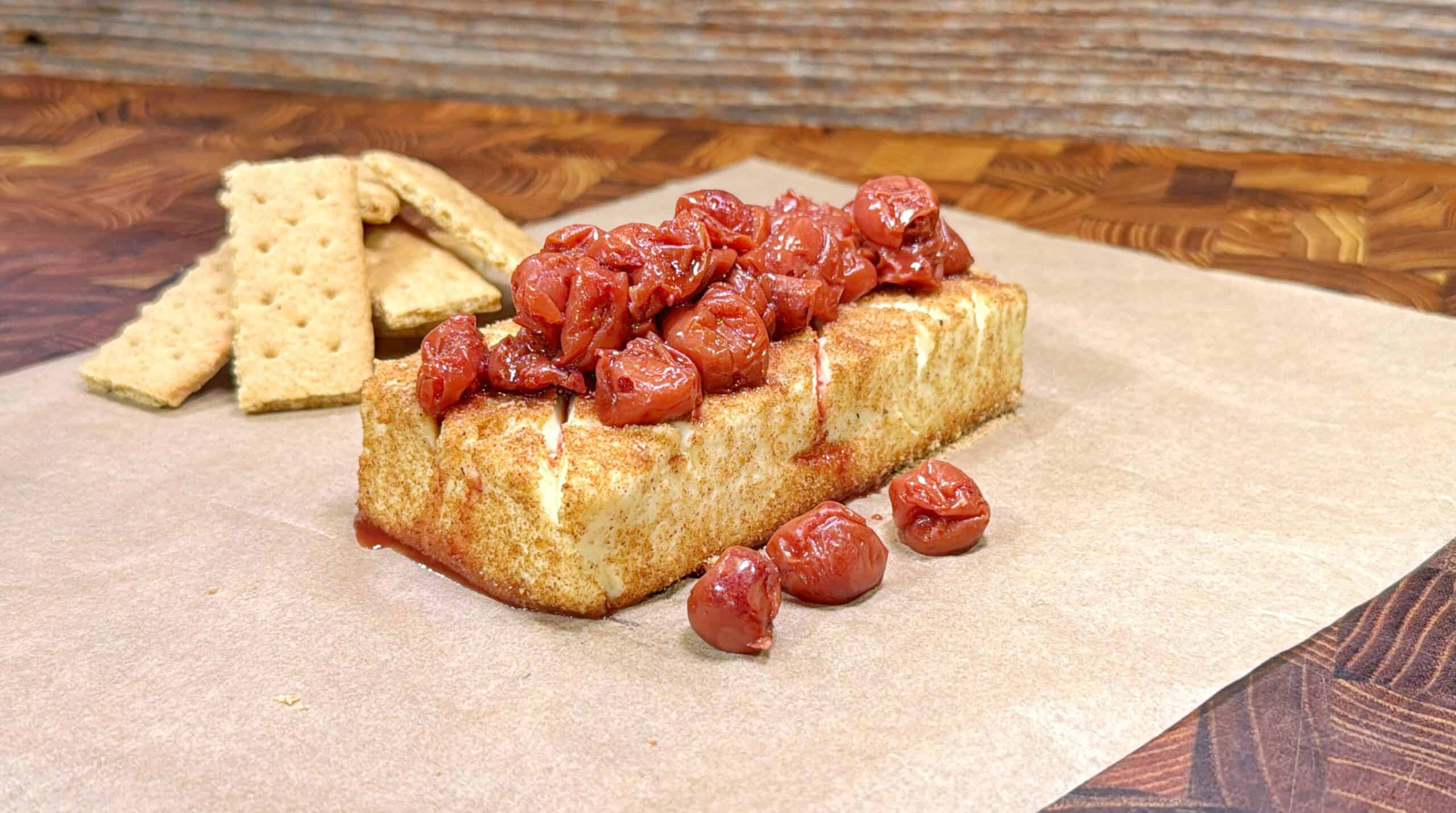 A slice of cheesecake topped with cherries sits on parchment paper, with several graham crackers stacked in the background on a wooden surface.