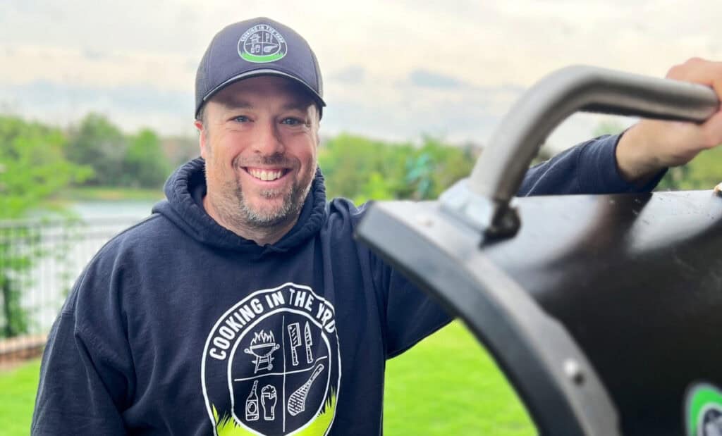 A smiling man in a black hoodie and cap stands outdoors by a barbecue grill. The hoodie reads COOKING IN THE YOO. Green grass and trees are visible in the background, setting the scene for an Oh That’s Good cookout.