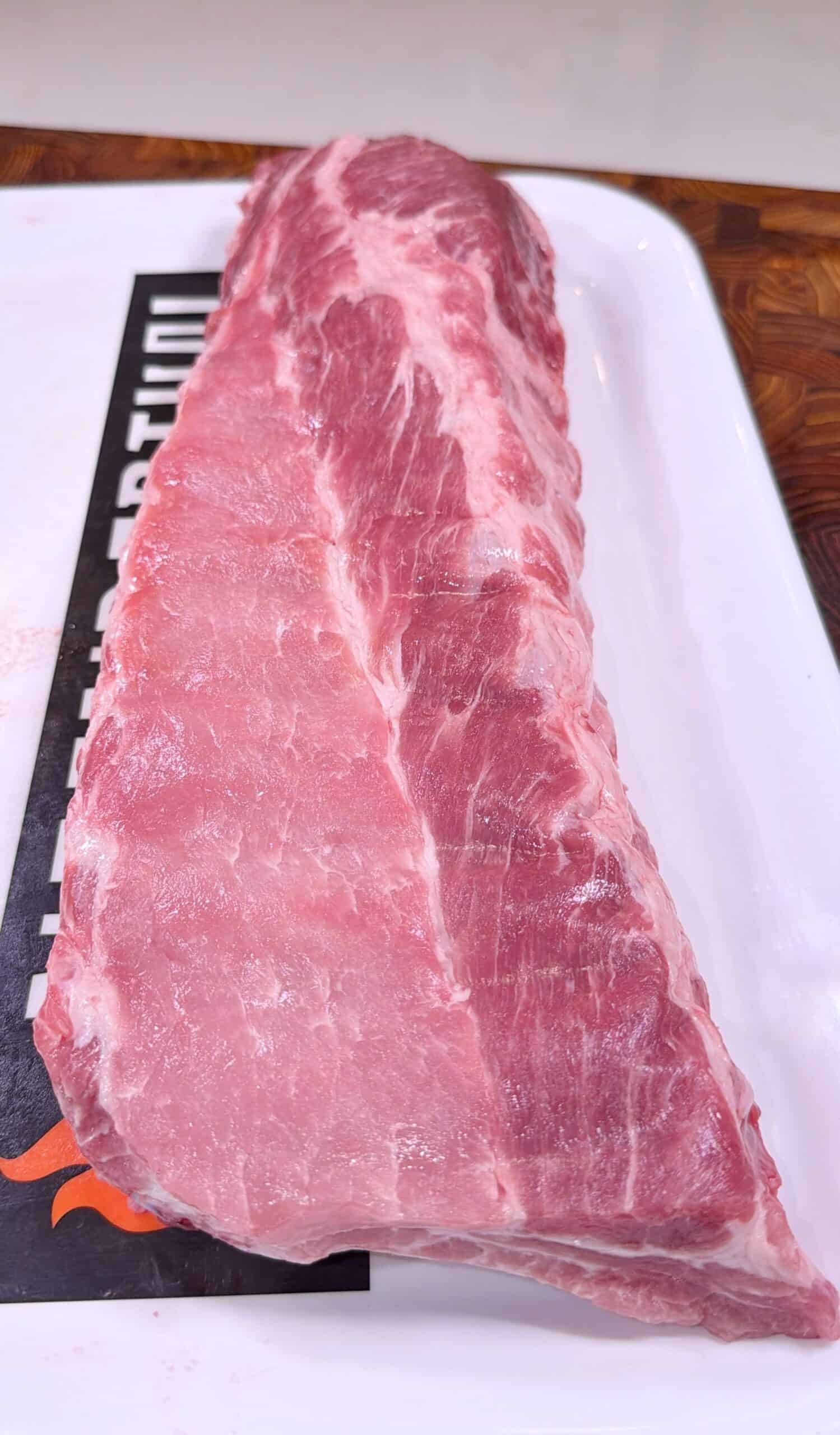 A large, raw slab of pork ribs sits on a white rectangular tray, displaying marbled pink meat and thin layers of fat. The tray is set on a wooden surface.