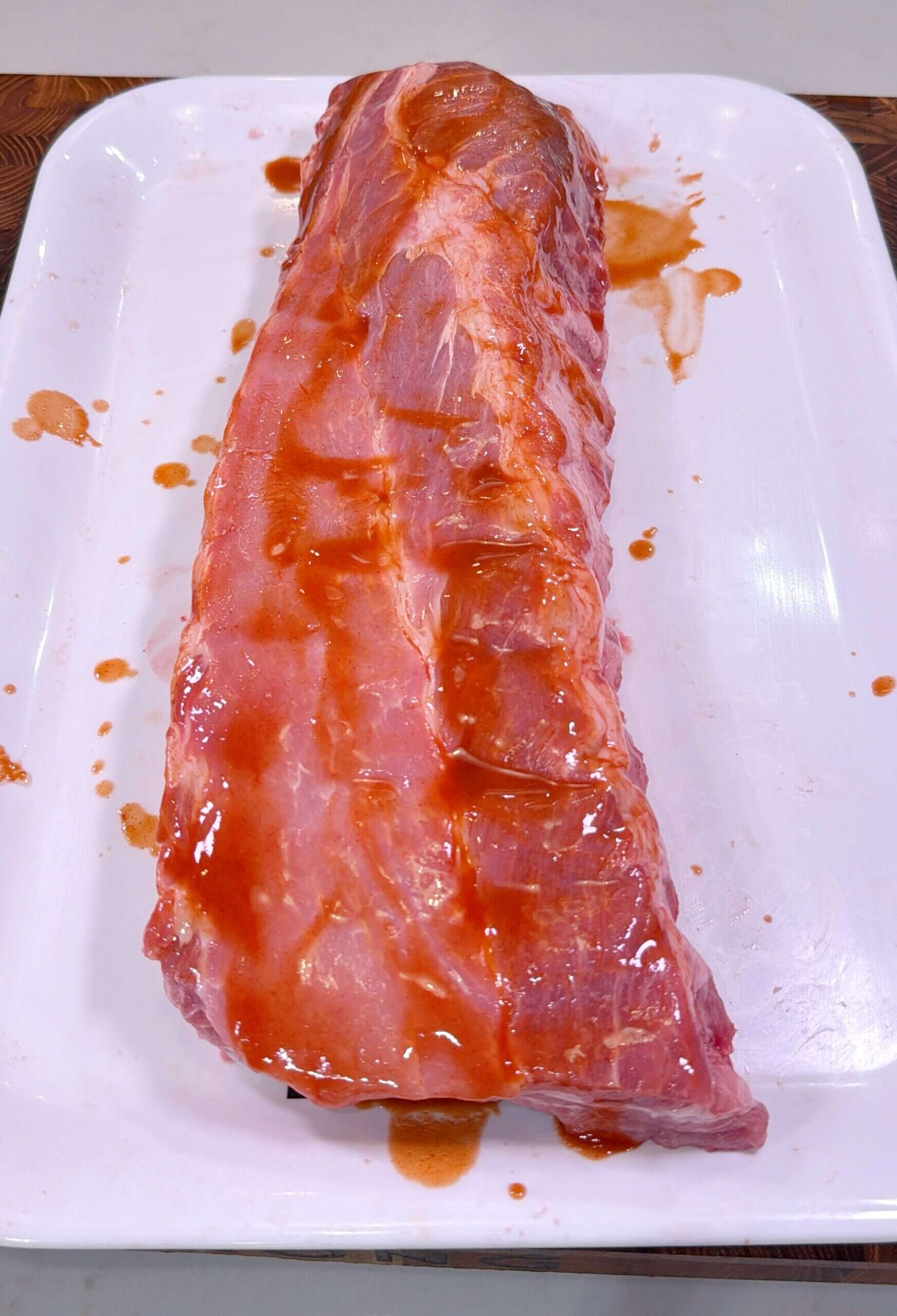 A raw rack of pork ribs covered with a layer of reddish marinade or sauce, placed on a white rectangular tray.