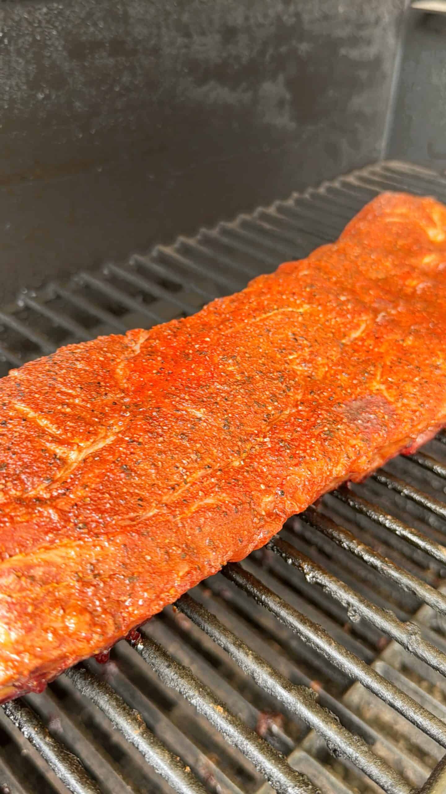 A rack of seasoned ribs with a reddish spice rub is cooking on a grill, with grill marks starting to form on the meat. The background shows the dark, metal grates of the grill.