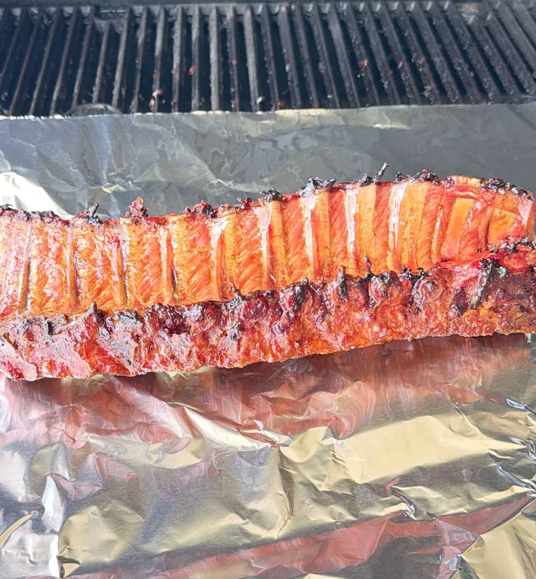 Barbecued rack of ribs with a crispy, caramelized exterior rests on aluminum foil with a grill grate visible in the background.