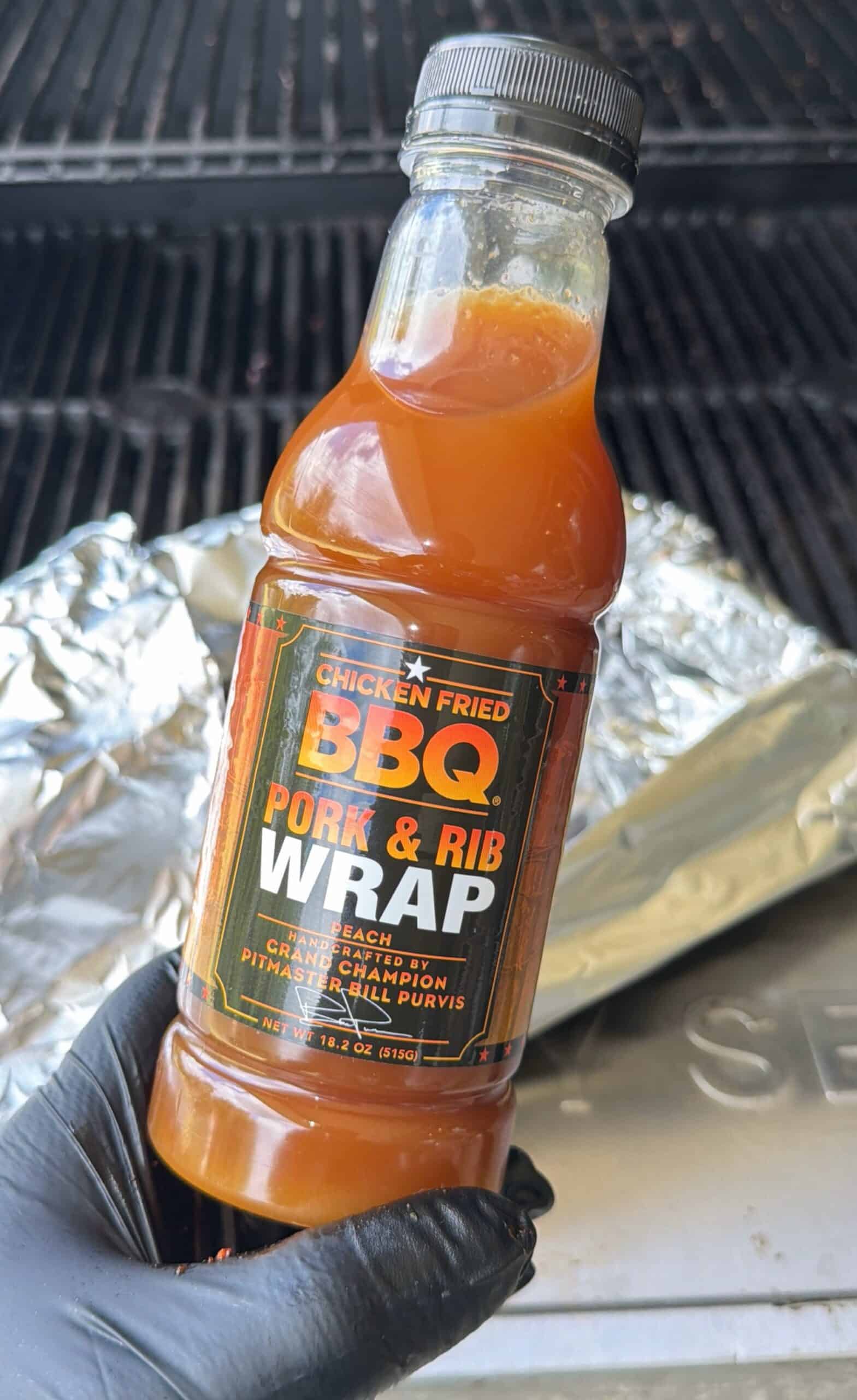 A gloved hand holds a bottle of Chicken Fried BBQ Pork & Rib Wrap sauce in front of a foil-wrapped item on a grill. The sauce bottle is partially filled with reddish-orange liquid.