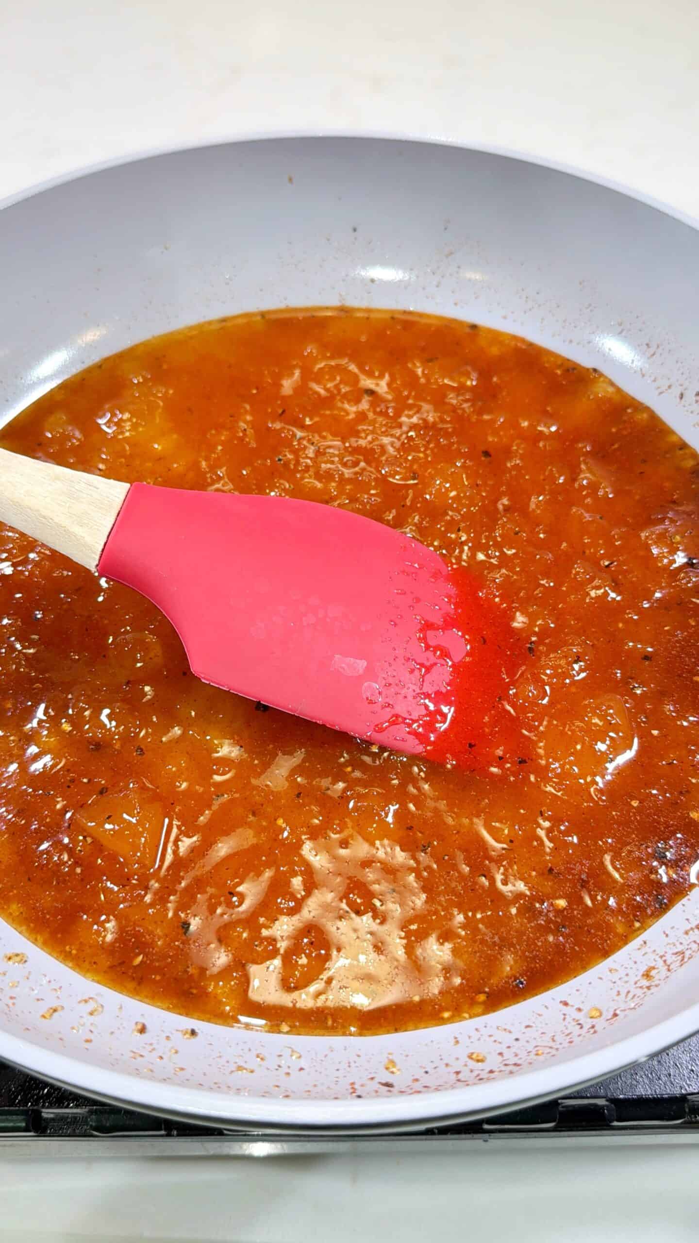 A red silicone spatula stirs a pan of simmering red-orange sauce or stew, with visible oil and spices on the surface. The pan has a light-colored interior and is on a stovetop.