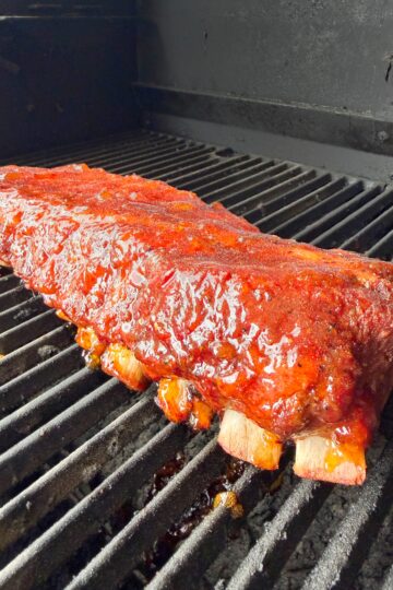 A rack of glazed, barbecued ribs cooks on a black grill, with a shiny, caramelized sauce coating the meat and grill marks visible underneath.
