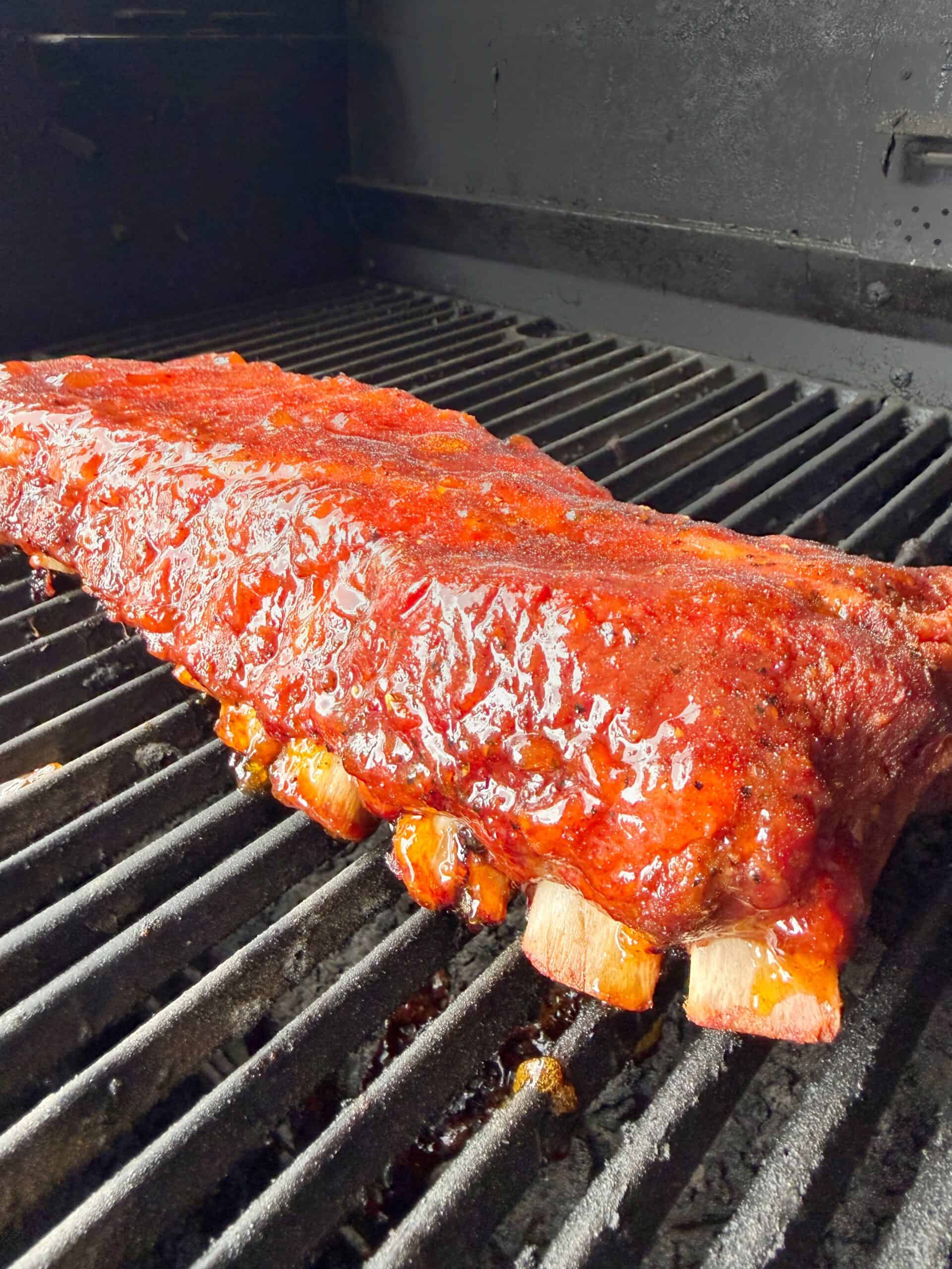 A rack of glazed, barbecued ribs cooks on a black grill, with a shiny, caramelized sauce coating the meat and grill marks visible underneath.