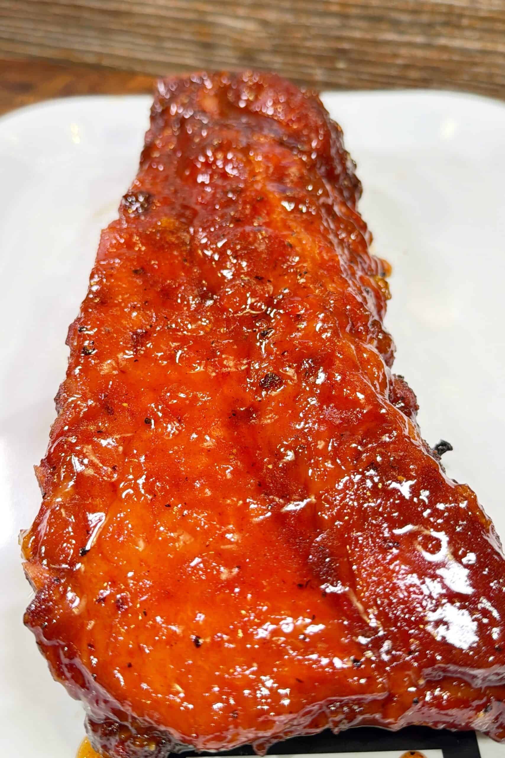 A close-up of a rack of glazed, barbecued ribs on a white plate, with a shiny, caramelized sauce coating the meat.
