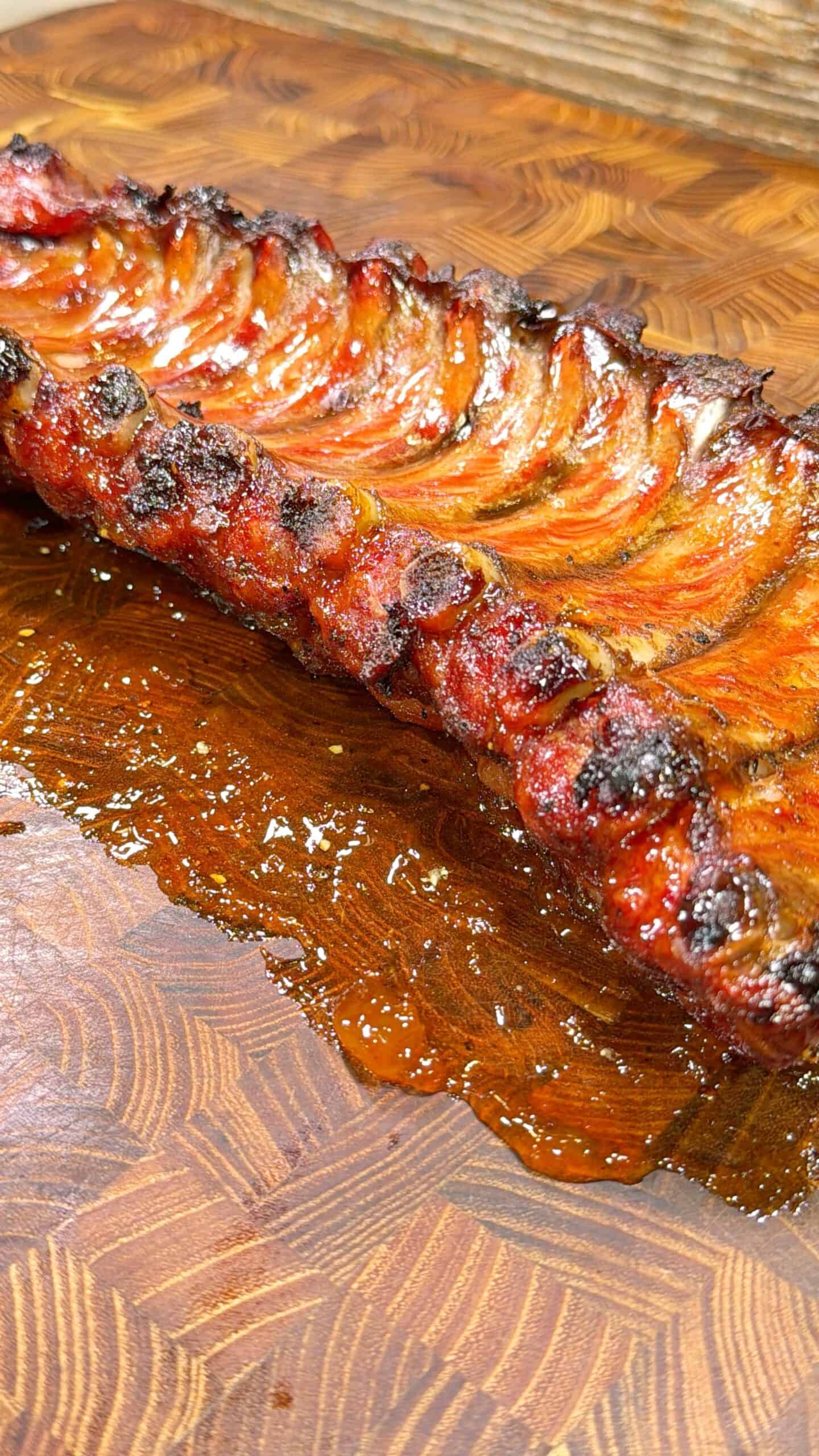 A rack of grilled, caramelized pork ribs rests on a wooden cutting board, glistening with sauce and juices, displaying charred edges and a rich, golden-brown color.
