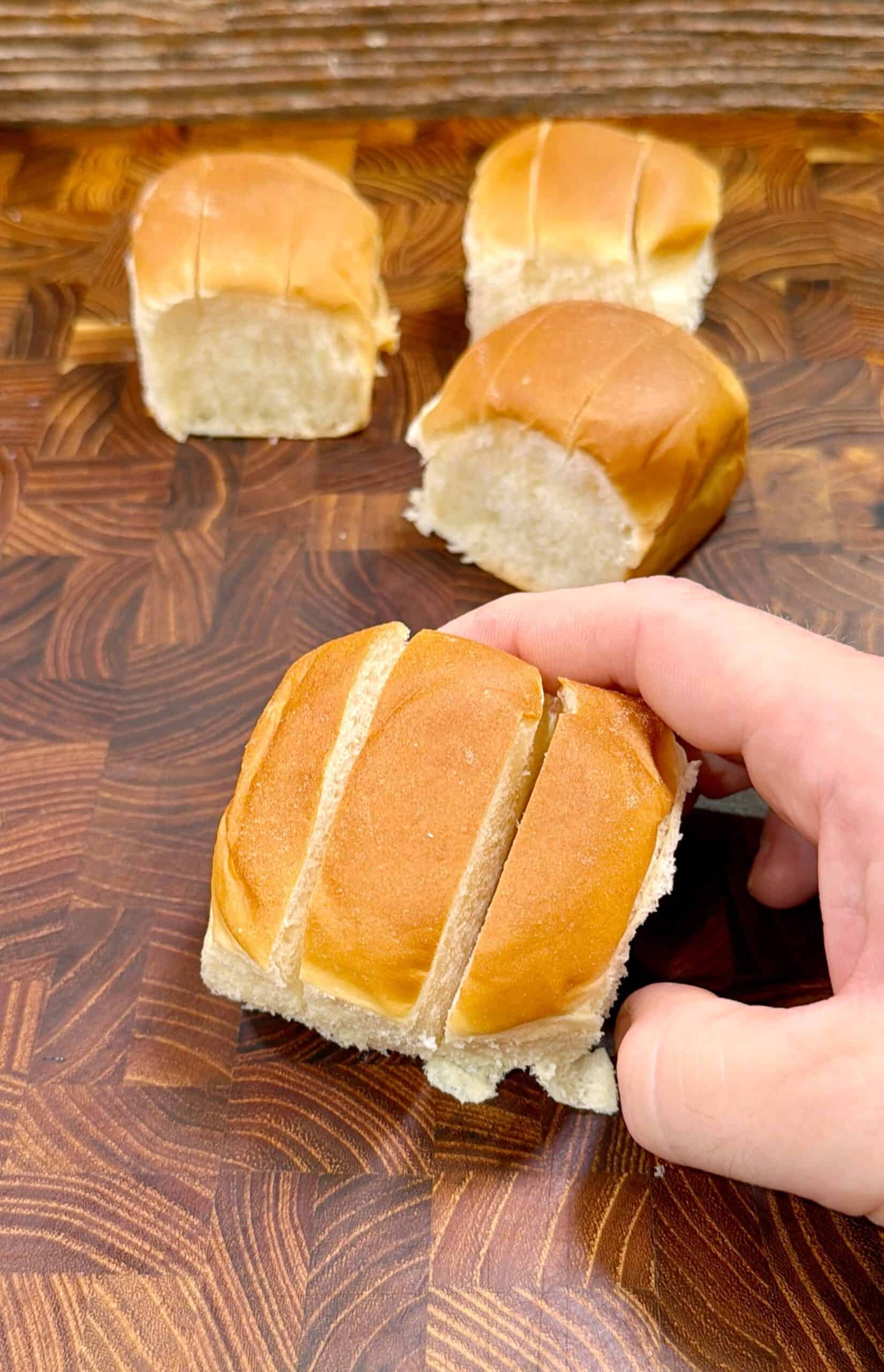 A hand holding a soft dinner roll sliced into three pieces, with three more sliced rolls resting on a wooden cutting board in the background.