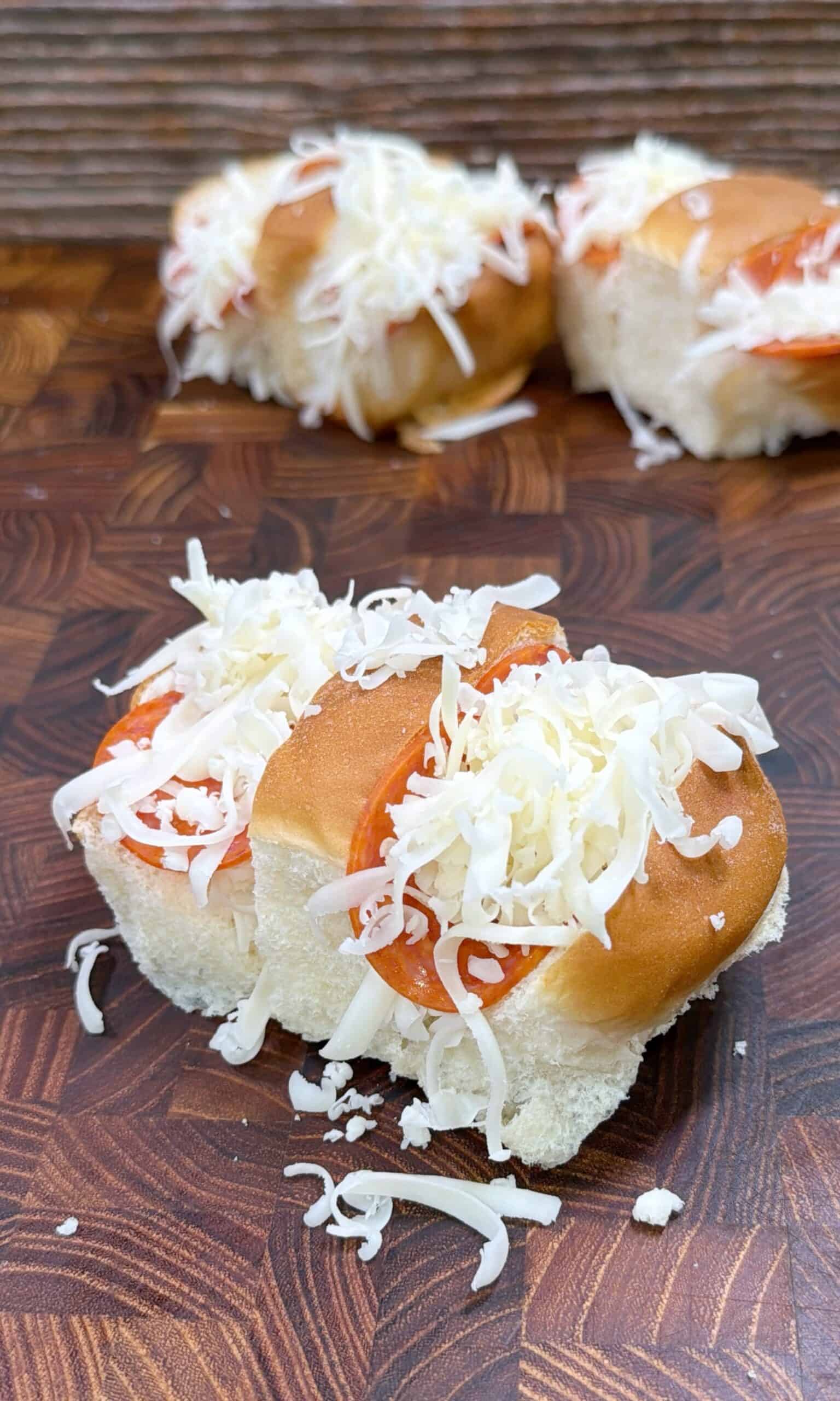 A close-up of soft sandwich buns filled with slices of pepperoni and topped with a generous amount of shredded mozzarella cheese, set on a wooden surface. Another similar bun is visible in the background.