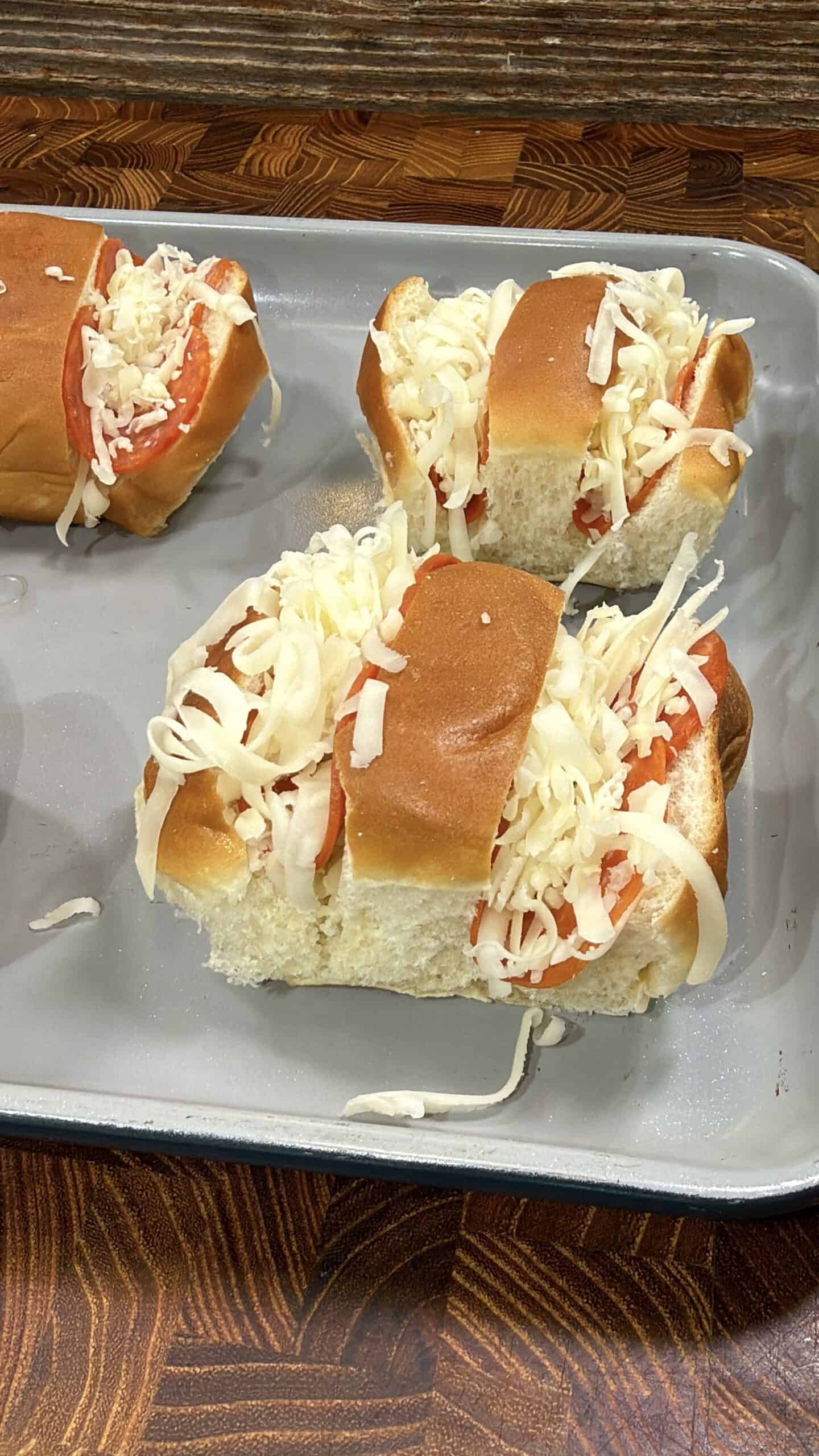 Three sandwich buns in a baking tray filled with sliced tomatoes and topped with shredded cheese, resting on a wooden surface.