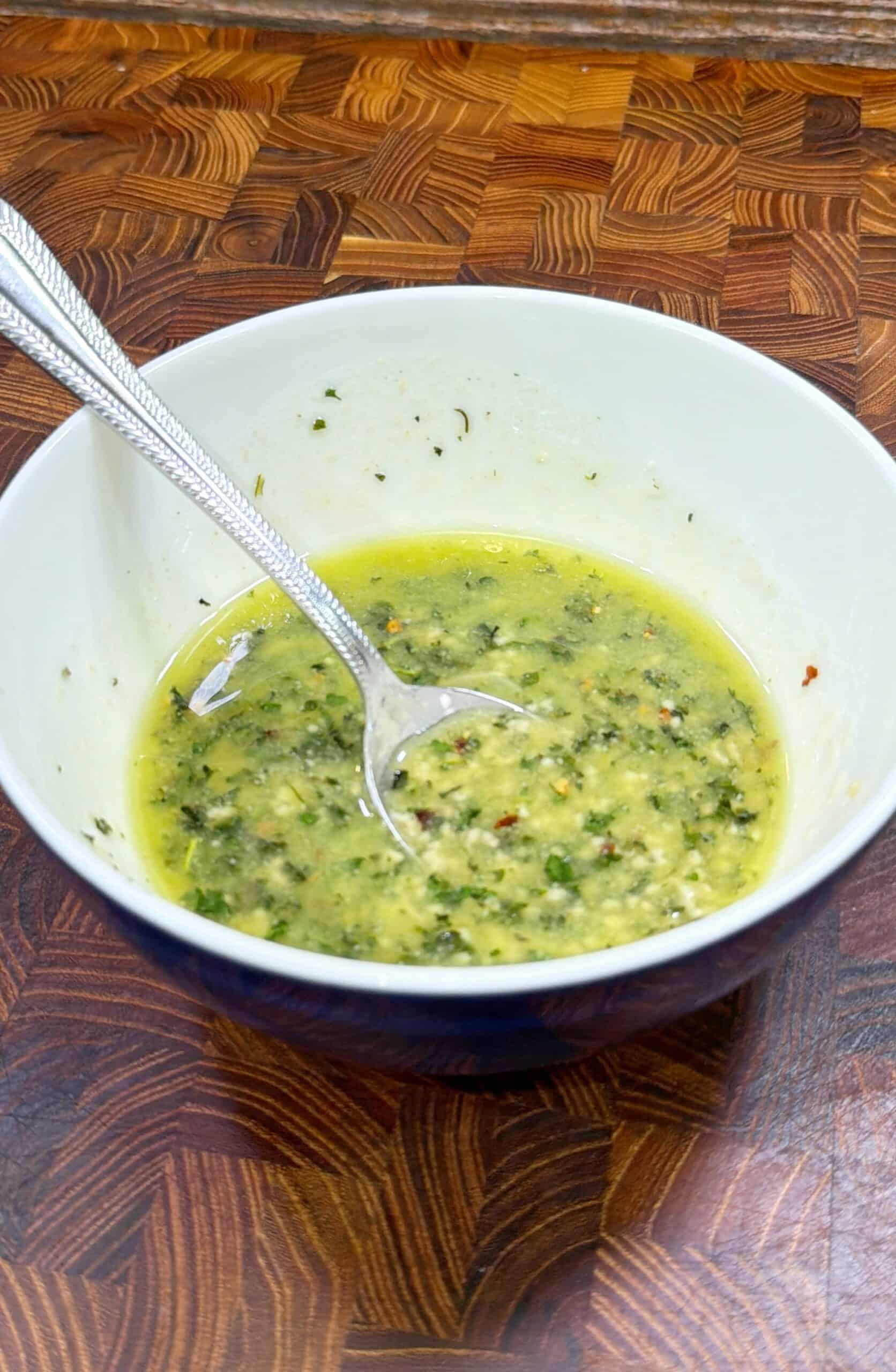 A white bowl with a metal spoon contains a yellow-green sauce with visible herbs and seasonings, sitting on a wooden patterned countertop.