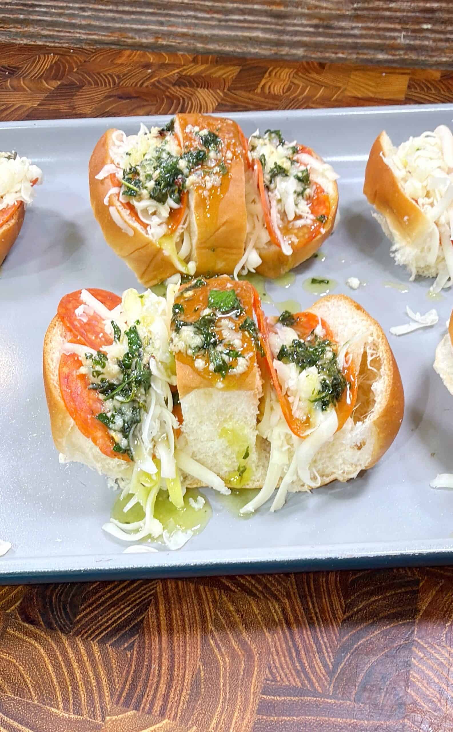 Mini sandwiches filled with sliced pepperoni, shredded cheese, and topped with a green herb sauce, served on a rectangular gray plate on a wooden table.